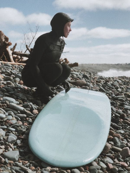 Person in a full black wetsuit crouching on a rocky beach next to a light blue surfboard, looking out toward the water.