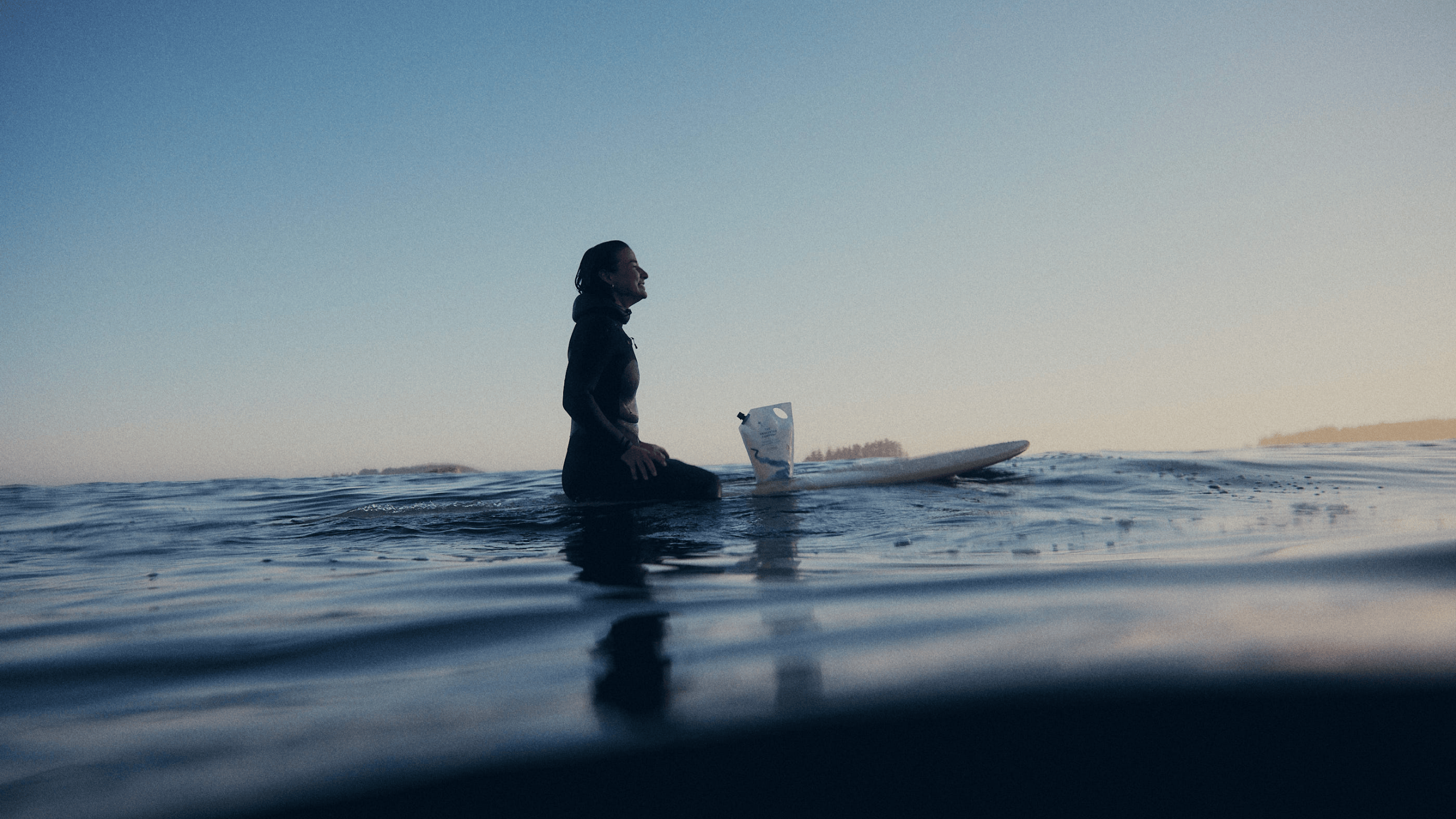 A surfer in a wetsuit sitting on a surfboard in calm ocean water at dusk, with a The Unscented Company refill pouch resting on the board beside them.