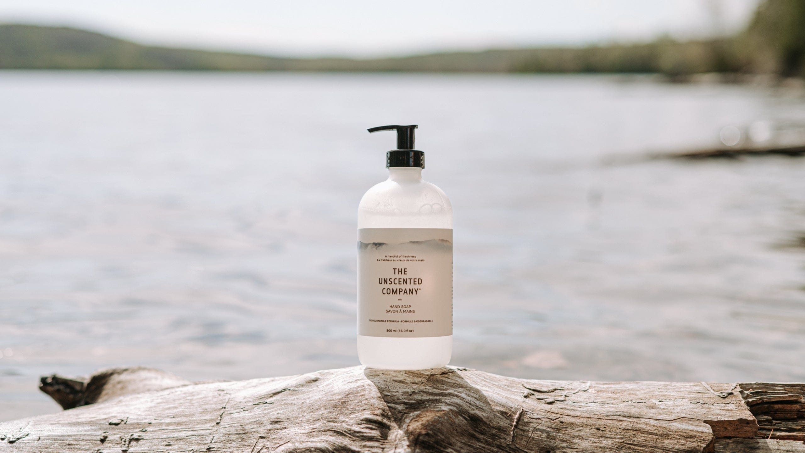 A bottle of The Unscented Company hand soap placed on a piece of driftwood by a lake, with calm water and distant forested hills in the background.