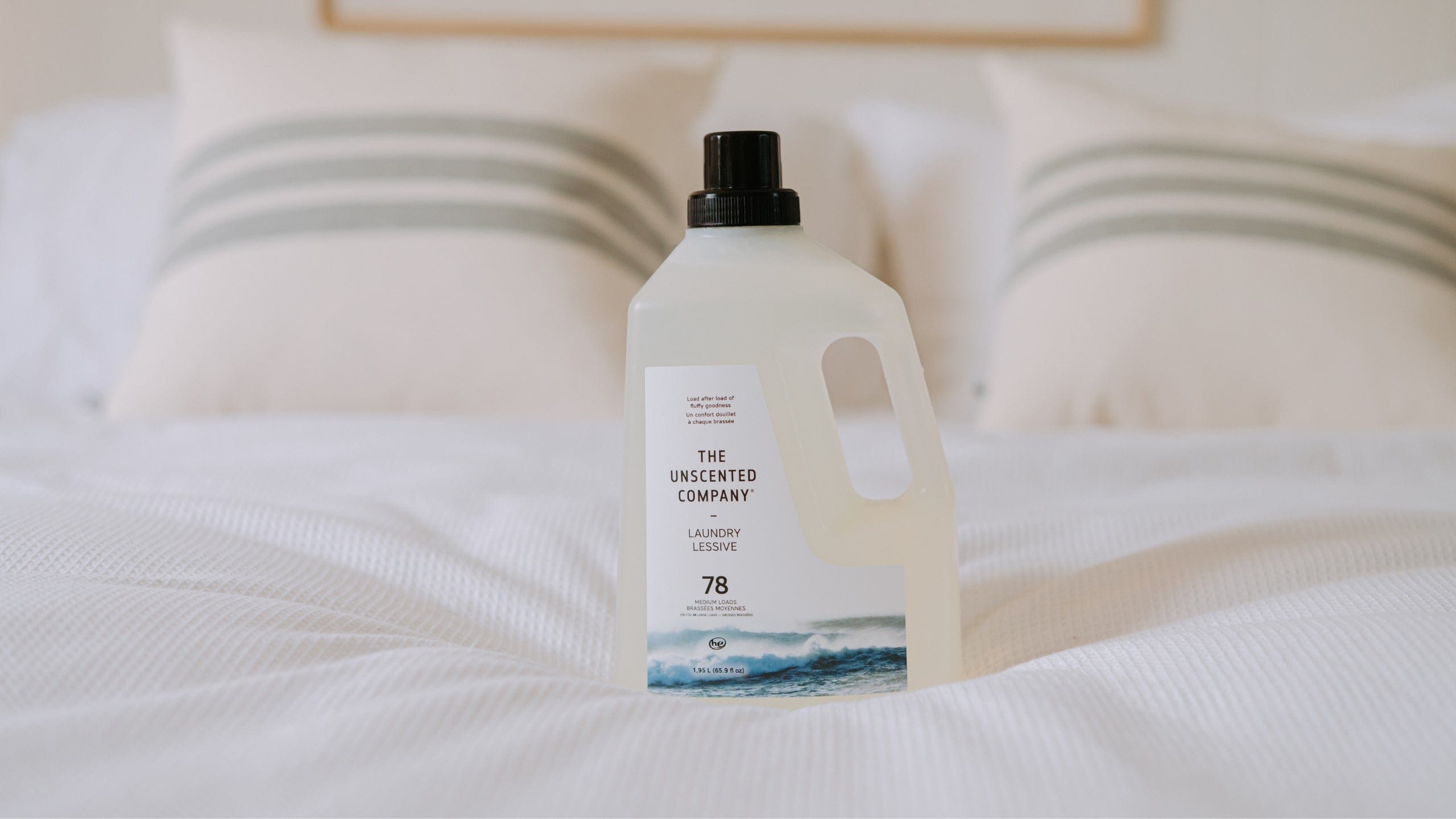 The Unscented Company liquid laundry detergent bottle placed on a white bed, flanked by two cream-colored pillows with green stripes, in a bright and minimalist bedroom.