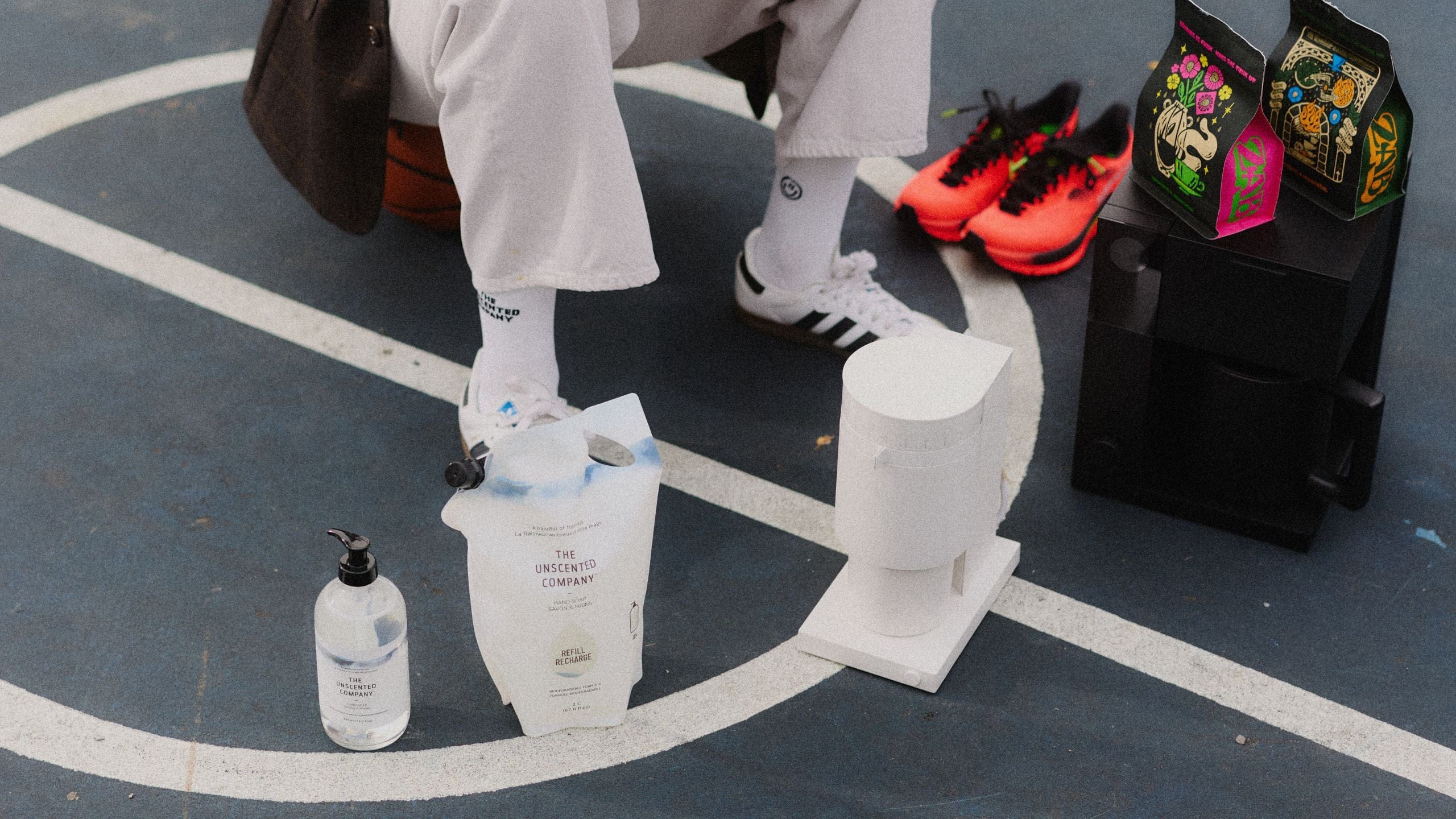 A person sitting on a basketball court with The Unscented Company products at their feet, including a hand soap refill pouch, a pump bottle, and a white dispenser, surrounded by accessories such as sports shoes and brightly designed coffee bags.