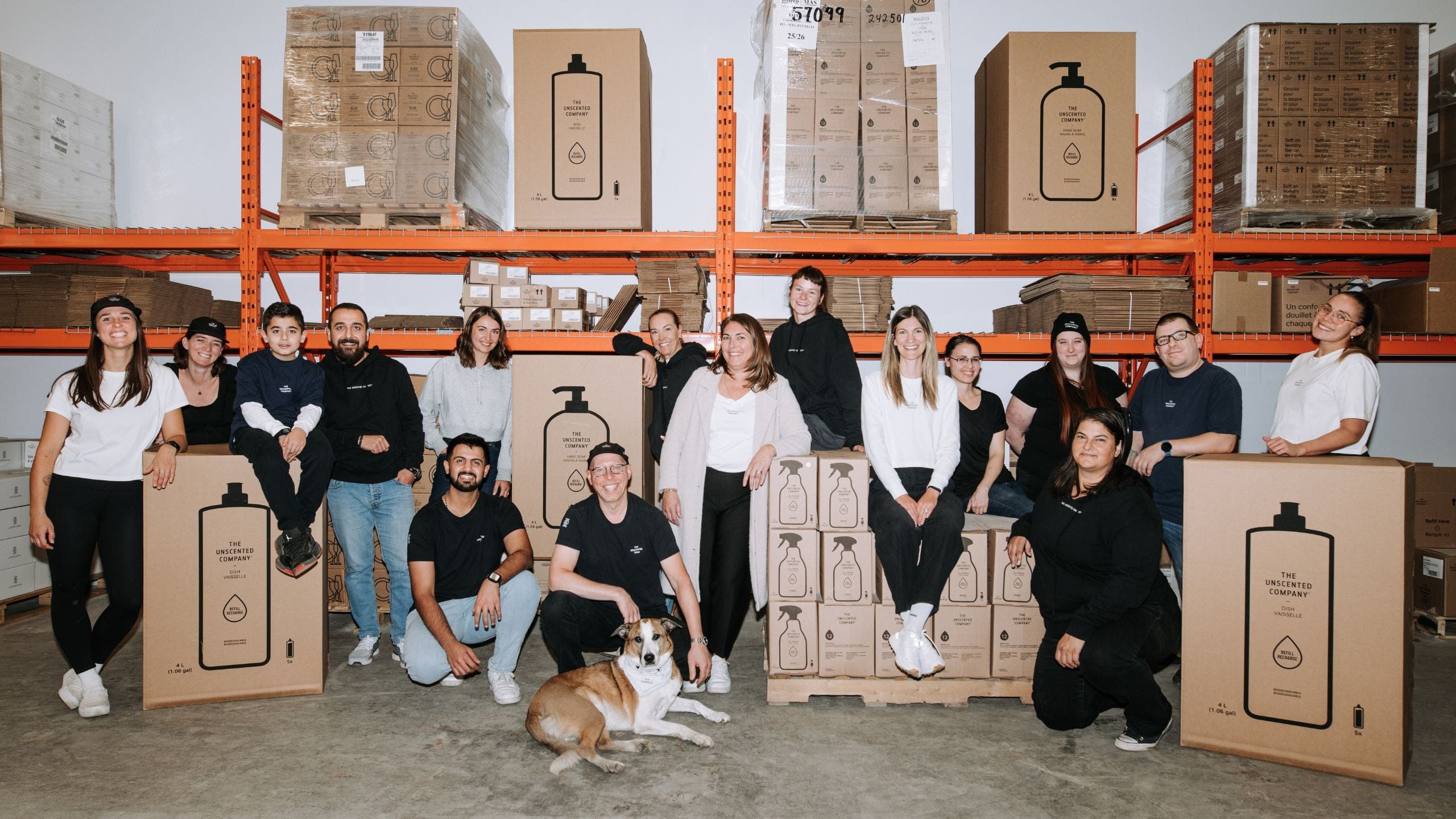 Group photo of The Unscented Company warehouse team posing in front of stacked pallets and orange shelving. Large cardboard boxes printed with refill product graphics are displayed. A dog lies at the front of the group.