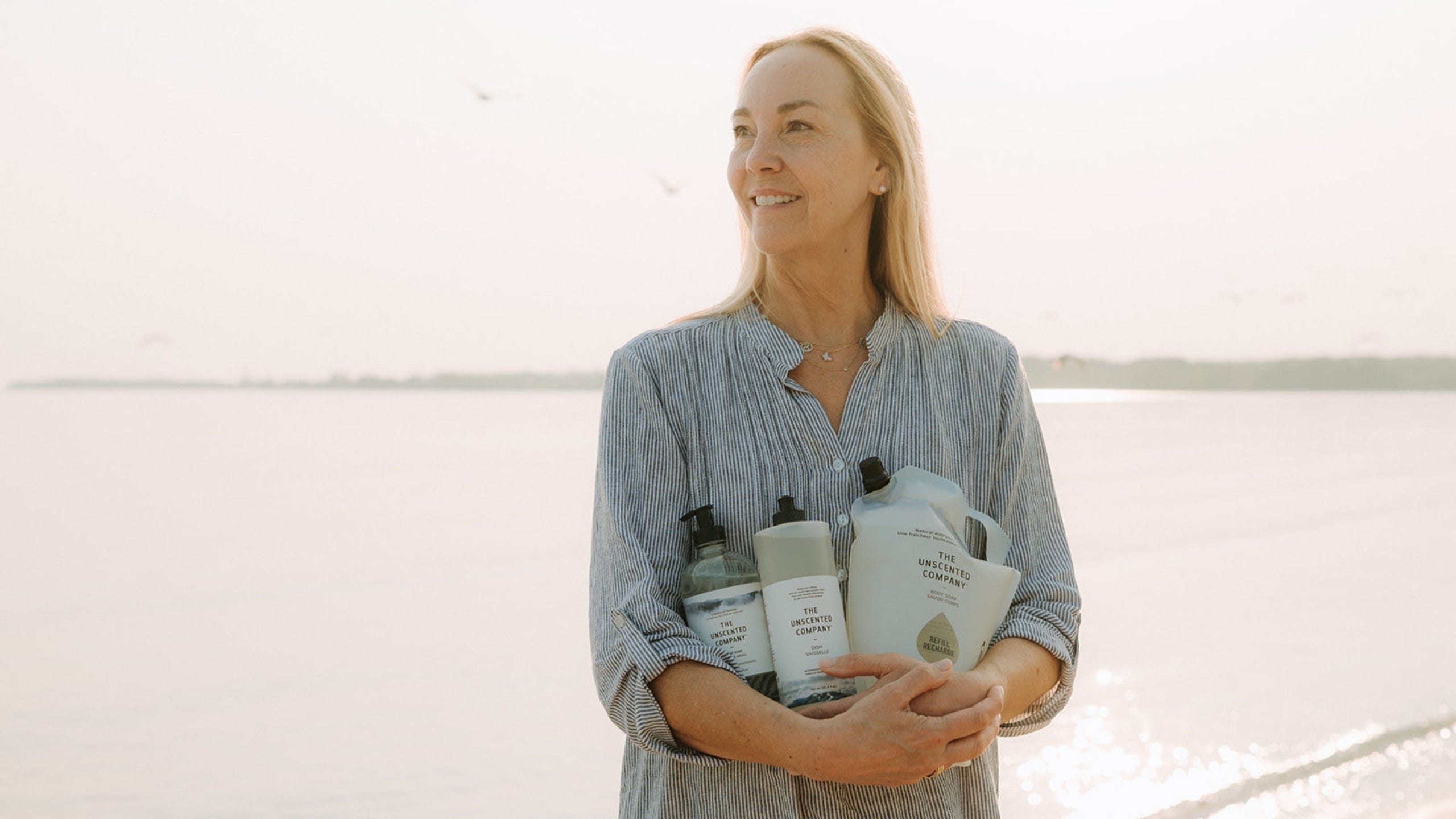 Smiling woman standing on a beach at sunrise, holding several The Unscented Company products in her arms.