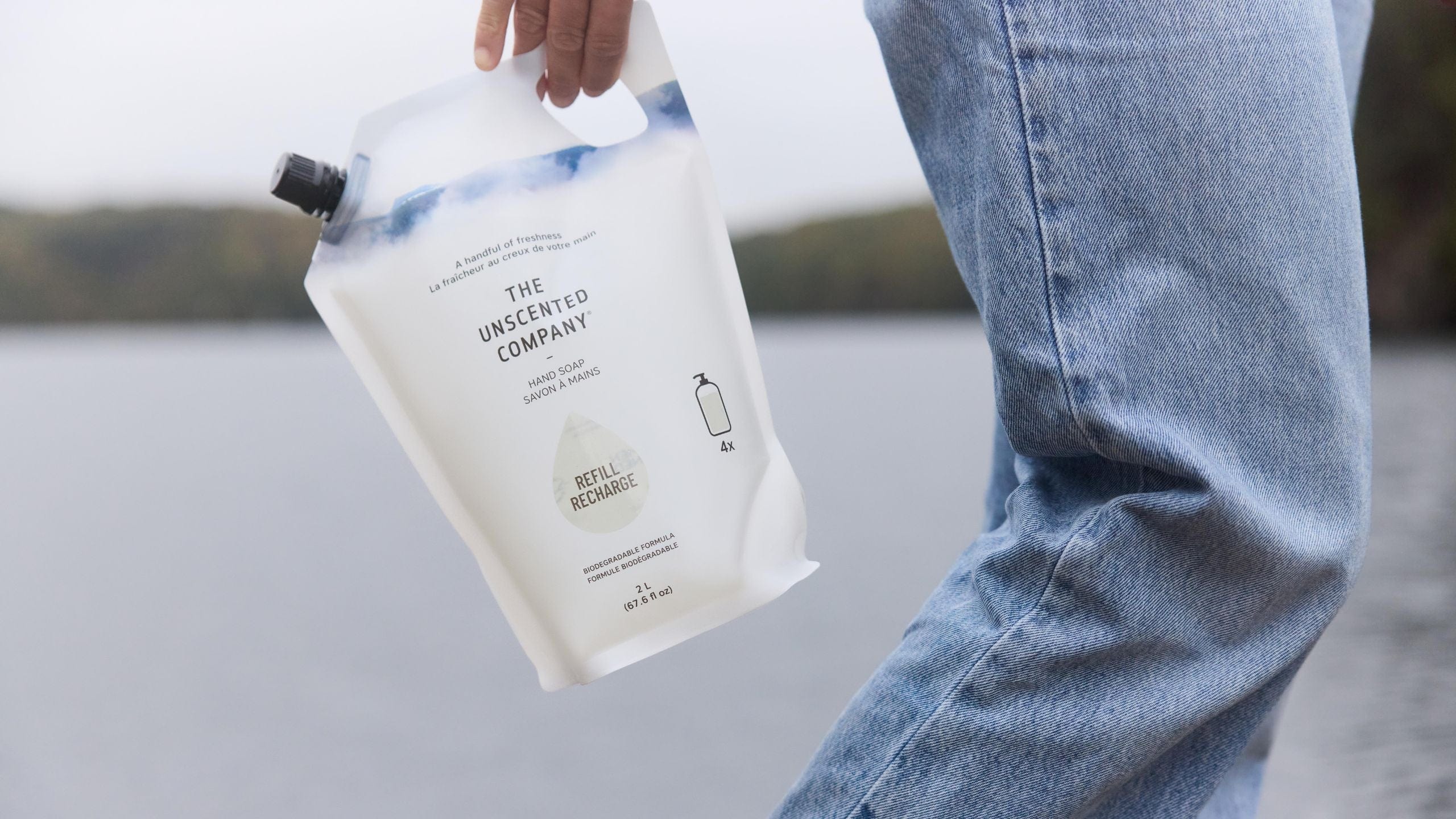 A hand holds a 2-liter refill pouch of The Unscented Company hand soap. The pouch reads “Refill Recharge.” A person in blue jeans stands beside a lake.