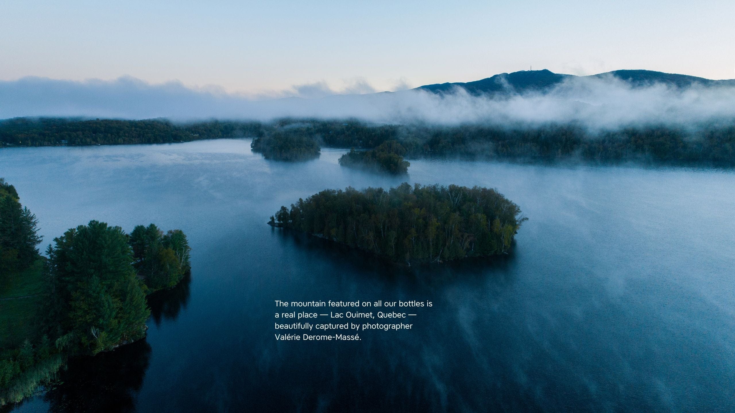 Aerial photo of Lac Ouimet, Quebec, showing forested islands, a calm lake, and a misty mountain in the background. On-screen text notes this mountain appears on all The Unscented Company bottles and was photographed by Valérie Derome-Massé.
