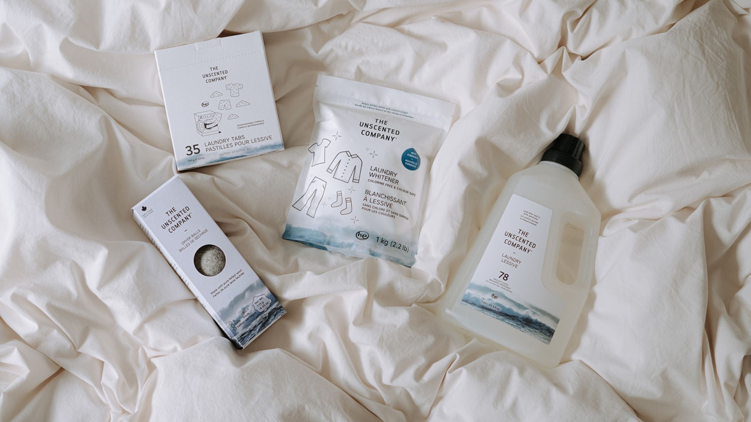 Four The Unscented Company laundry products placed on a white fabric surface: laundry tabs, laundry whitener, dryer balls, and liquid laundry detergent.