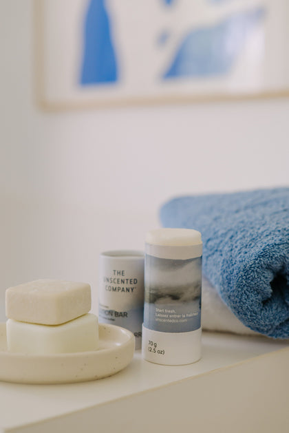 The Unscented Company Lotion Bar, Shampoo and Conditioner bars on a white ledge next to a blue towel in a minimalist bathroom.