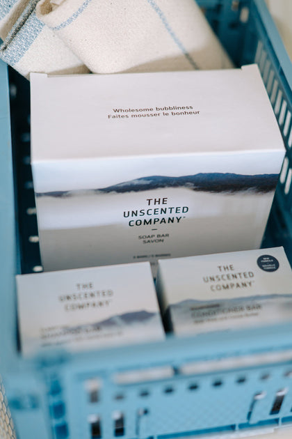 Close-up of The Unscented Company soap and conditioner bar boxes inside a blue storage crate with a beige towel.