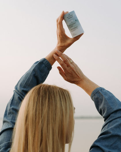 A woman with long blond hair, dressed in a denim shirt, holds up a deodorant stick from The Unscented Company with both hands against a soft pastel sky. The gesture suggests pride and appreciation for the product.