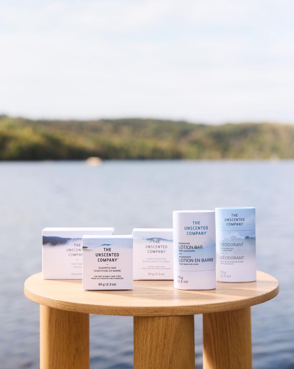 Various The Unscented Company personal care products — soap bar, shampoo bar, conditioner bar, lotion bar, and deodorant — arranged on a wooden table by a lake with trees in the background.