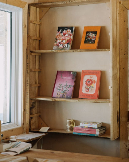 Wooden wall shelves displaying a colorful selection of flower and gardening books, including titles like 'Flora Mama', 'On Flowers', and 'Cut Flower Garden', beside a sunlit window in a cozy interior.