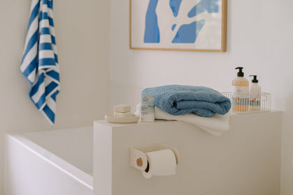 Bright bathroom featuring The Unscented Company products on a white bathtub ledge, with folded blue and white towels and a hanging toilet paper roll.