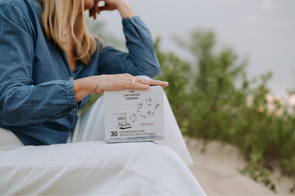 A woman sitting outdoors on a sandy surface, wearing a blue chambray shirt and white pants, rests her hand on a box of The Unscented Company dishwasher tabs. Green foliage and a soft sky frame the background.