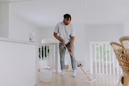 A man mops the floor of a bright, minimalist home using a reusable mop and a bucket filled with water. A pouch of The Unscented Company floor cleaner is visible in the background on the counter.
