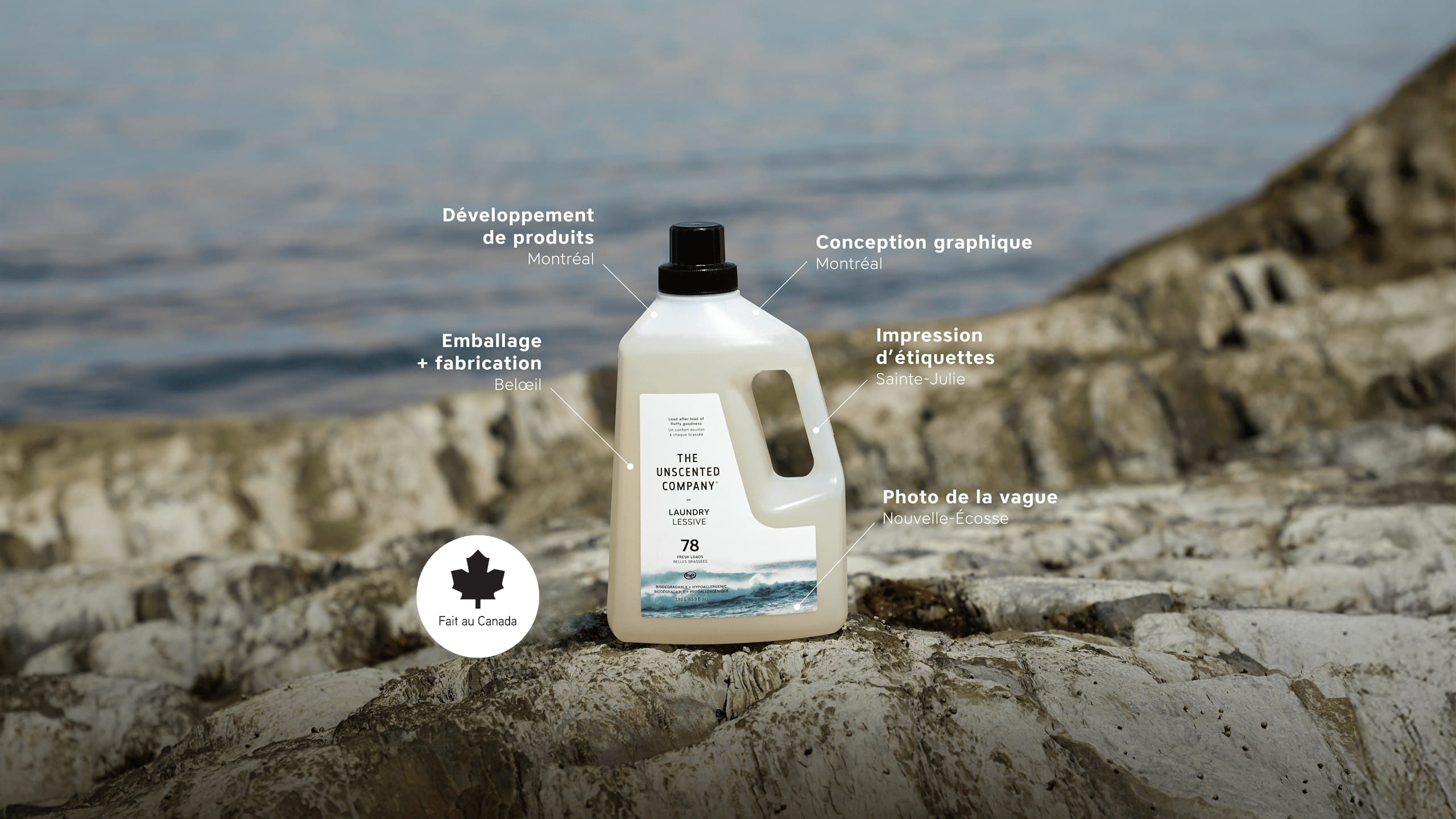 Bottle of The Unscented Company laundry detergent on rocky shoreline with water in the background. Text points to production steps: product developed in Montréal, graphic design in Montréal, packaging and manufacturing in Beloeil, label printing in Sainte-Julie, and wave photo taken in Nova Scotia. A “Fait au Canada” badge appears at the bottom left.