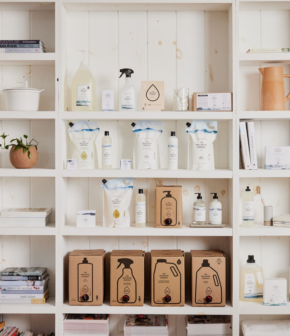 White wooden shelving filled with The Unscented Company products, including refill boxes, pouches, bottles, and soaps, surrounded by books and decor items.