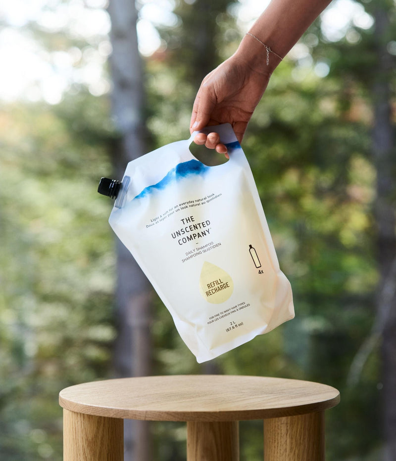 A hand holding a 2 L refill pouch of The Unscented Company daily shampoo above a small wooden table, with a forest in the background.