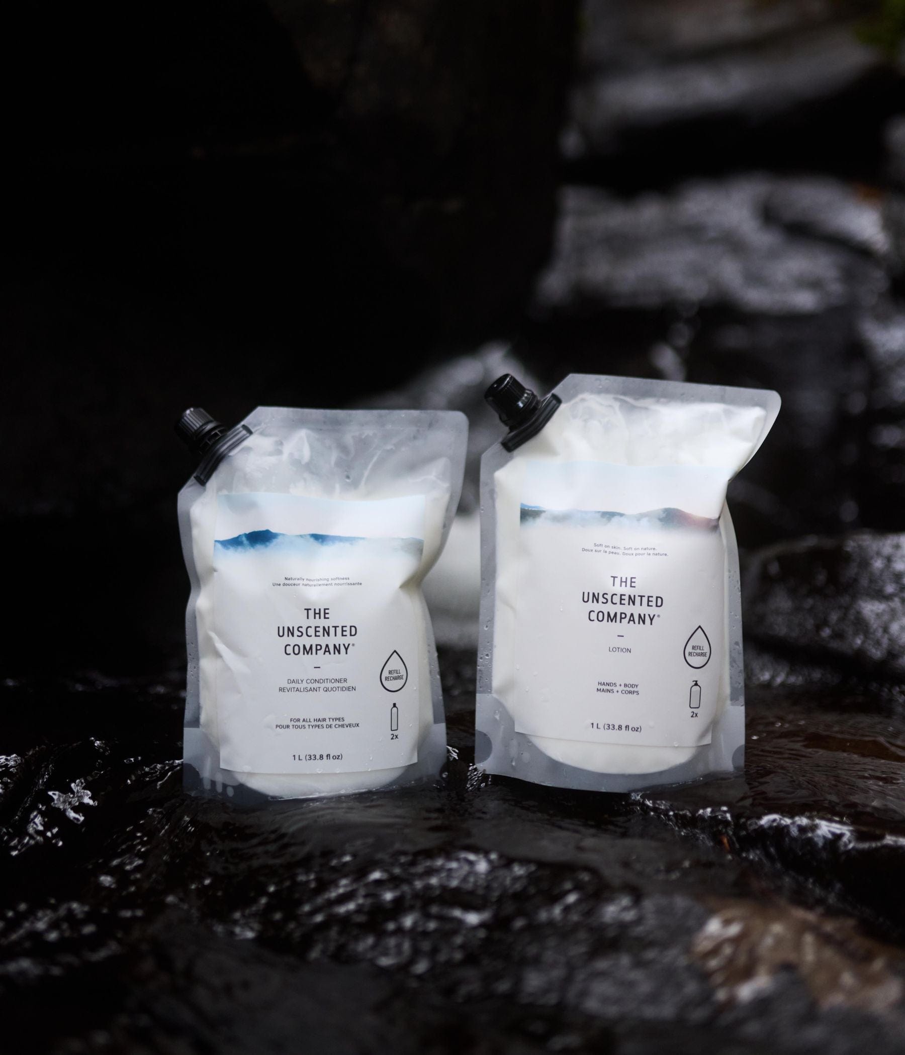 Two 1-liter refill pouches from The Unscented Company placed on wet black rocks. Left pouch is Daily Conditioner, right pouch is Lotion for hands and body.