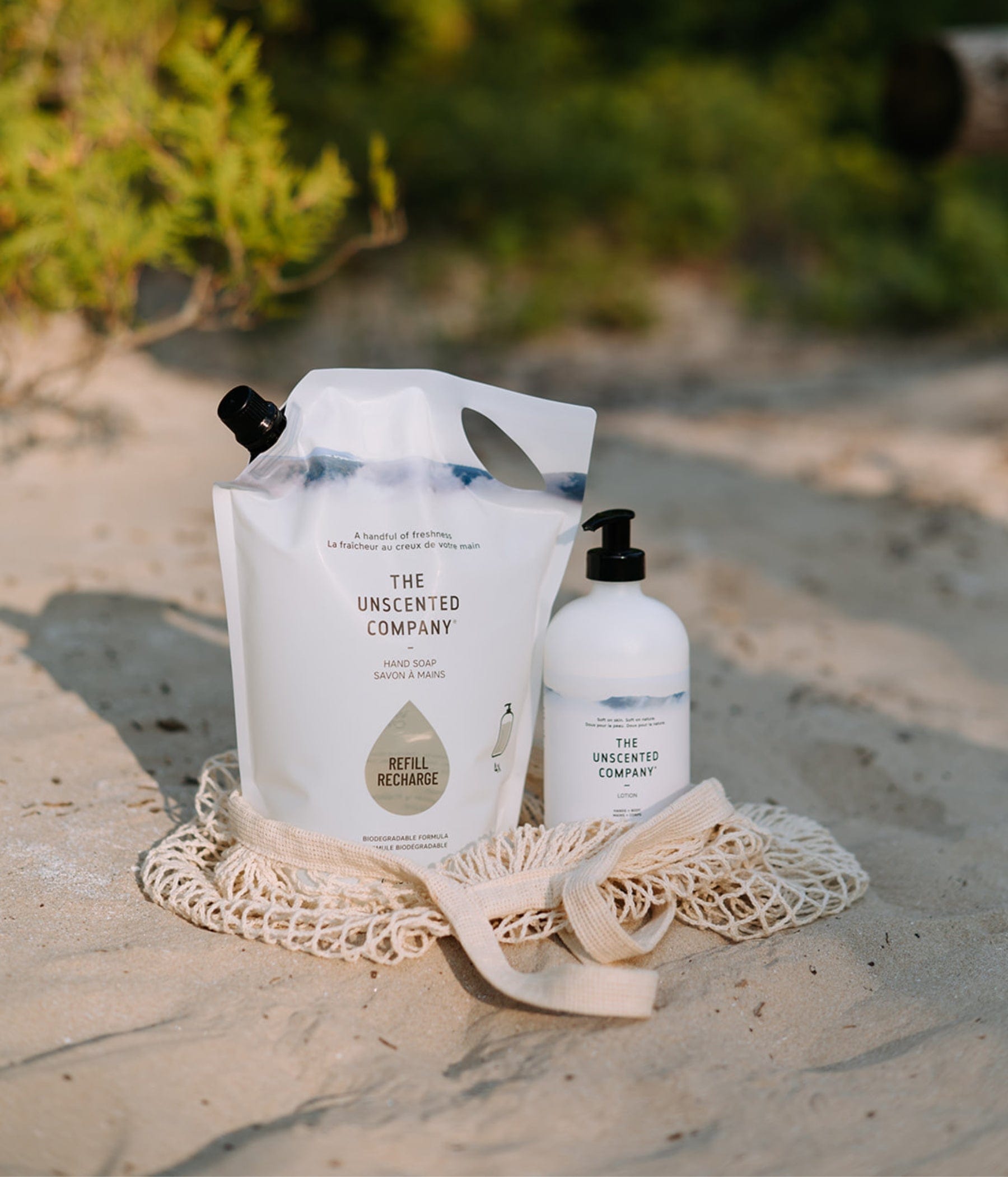 Two The Unscented Company products – a refill pouch of hand soap and a pump bottle – resting on a reusable net bag on the sand, surrounded by natural greenery.