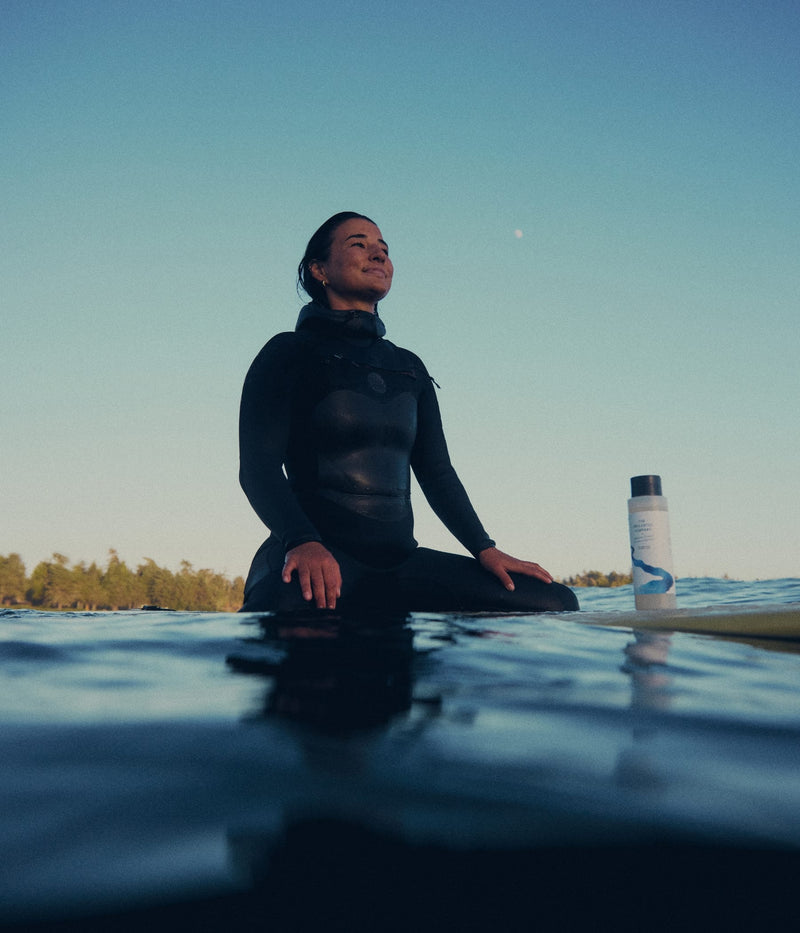 A surfer sitting on her surfboard in the water at sunset next to a bottle of hydrating shampoo from The Unscented Company.
