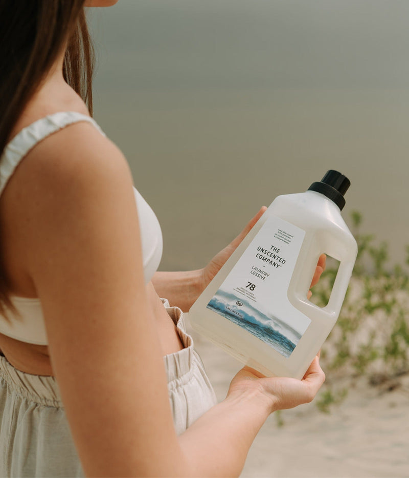 Woman holding a bottle of The Unscented Company laundry detergent on a sandy beach, near a calm lake.