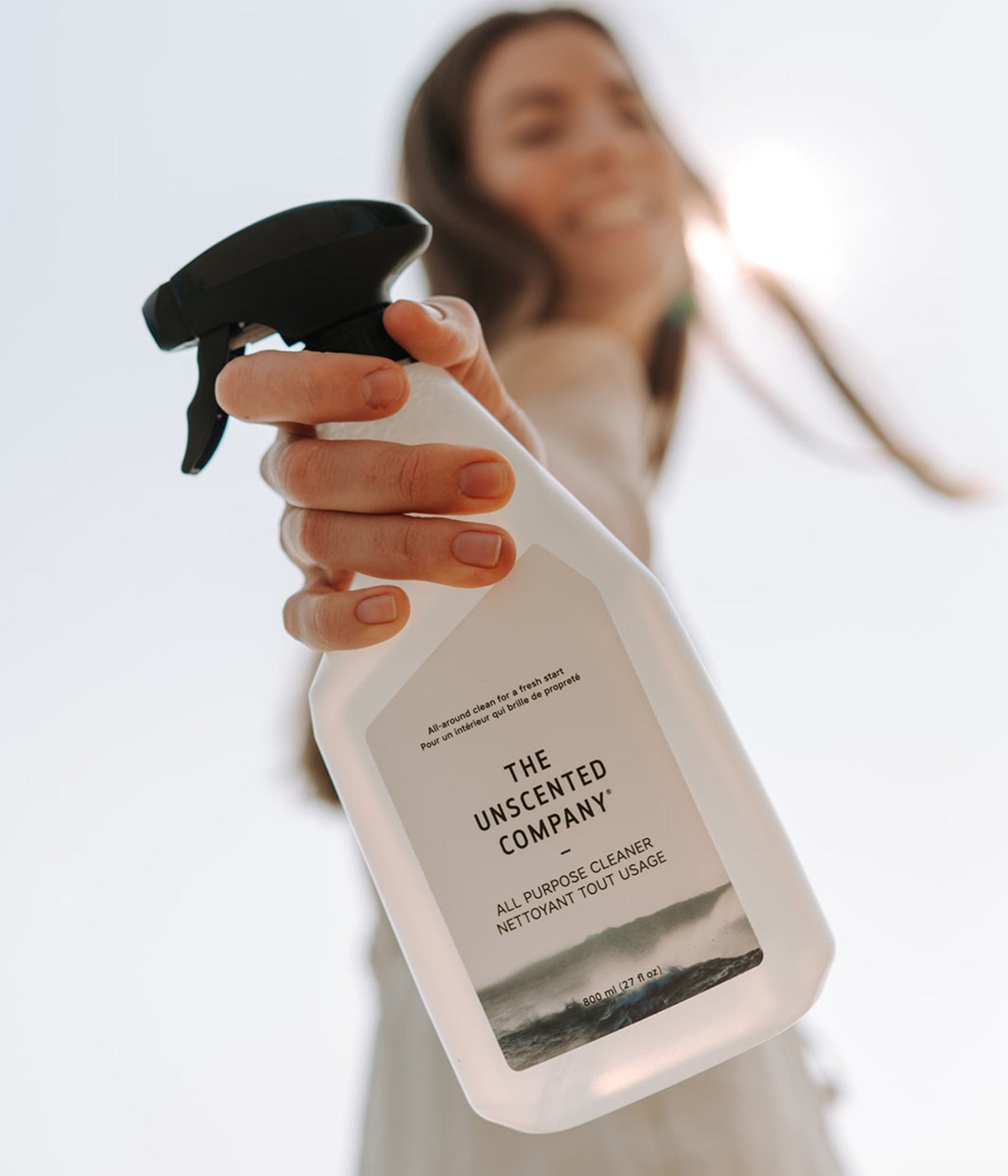 Smiling woman holding out a bottle of The Unscented Company all-purpose cleaner toward the camera, with natural backlight.