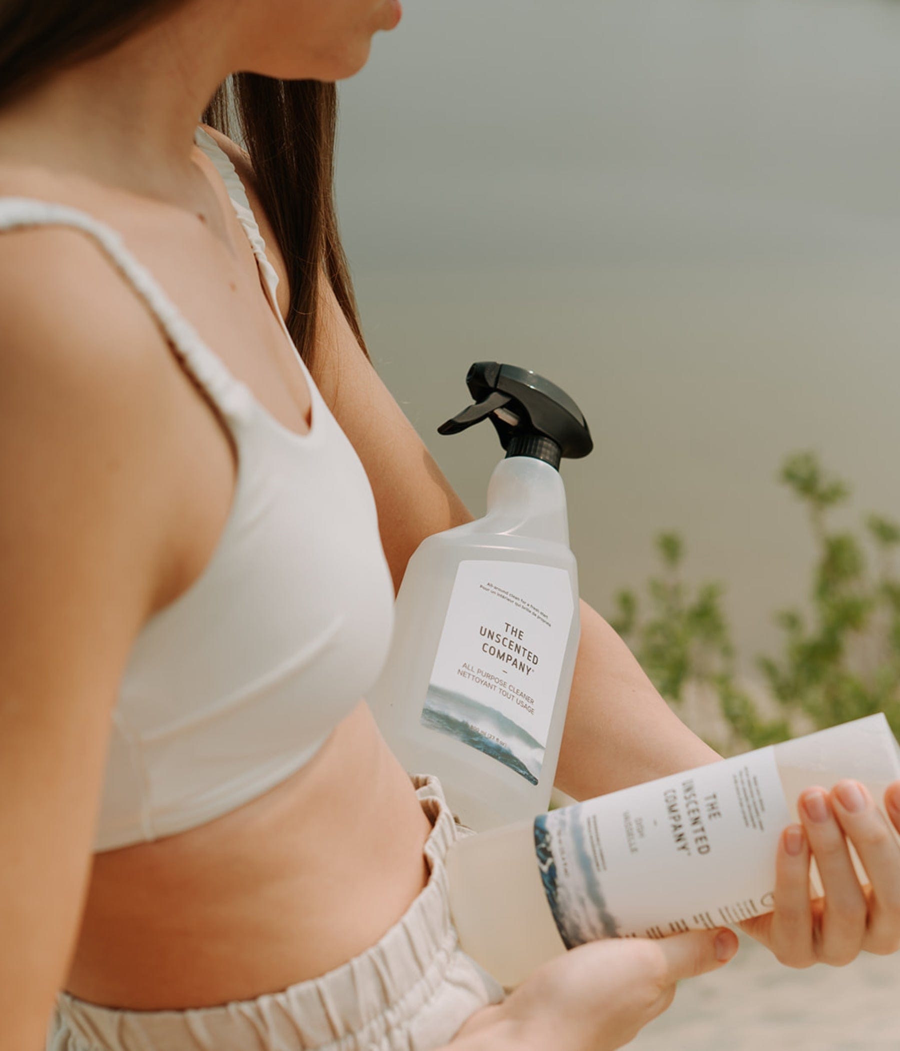 Woman in summer outfit holding a bottle of The Unscented Company all-purpose cleaner and soap, with a lake in the background.