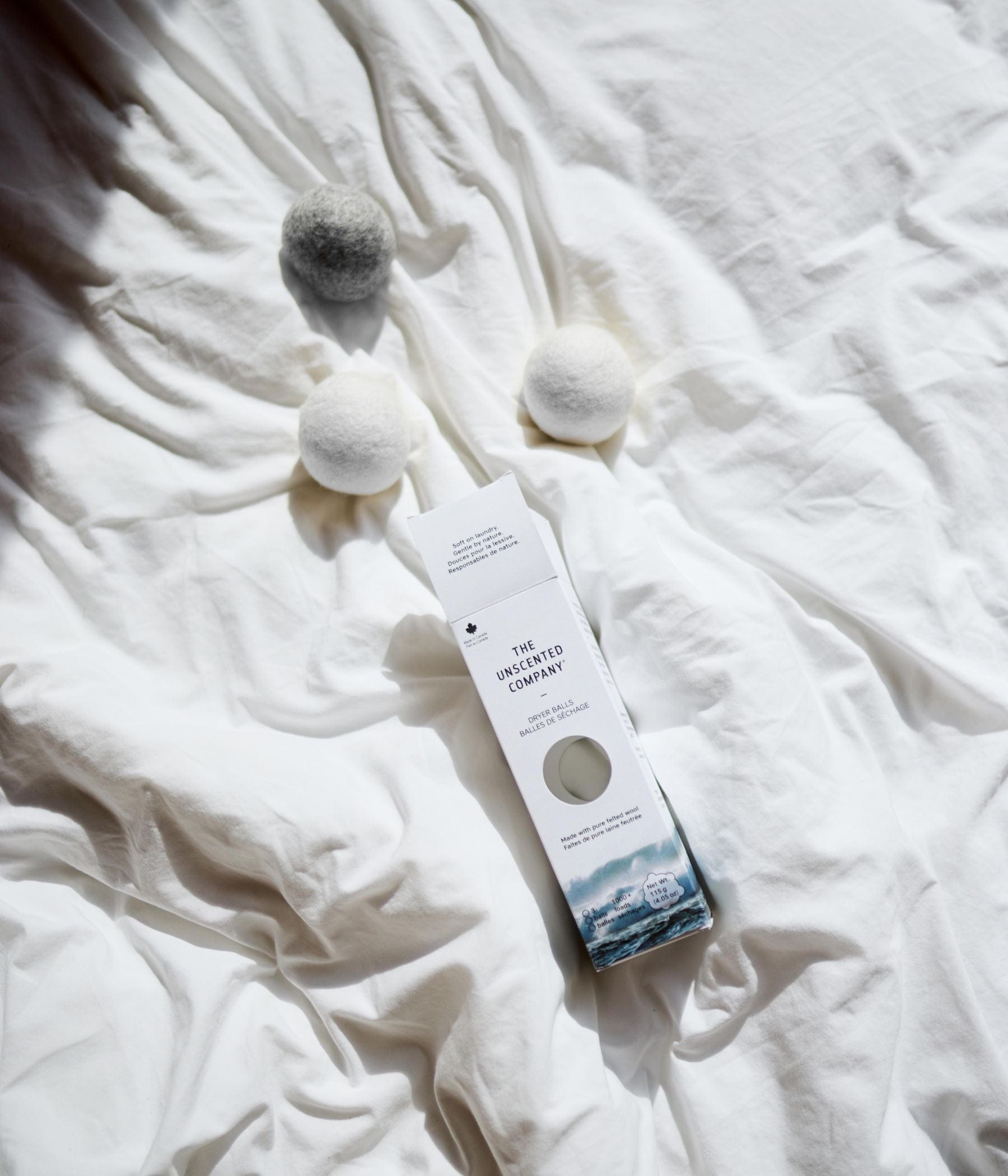 A box of The Unscented Company dryer balls and three wool dryer balls on a white bed sheet.