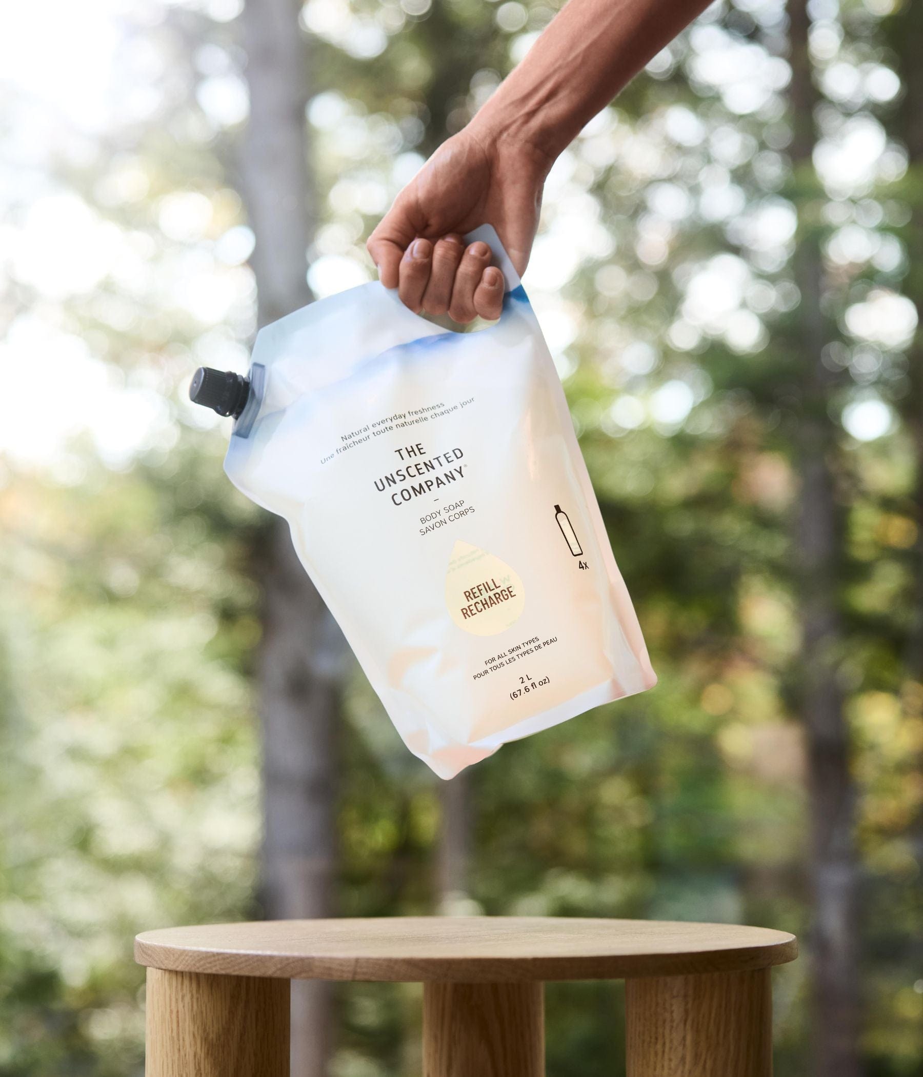 A hand holding a 2 L refill pouch of The Unscented Company daily shampoo above a small wooden table, with a forest in the background.