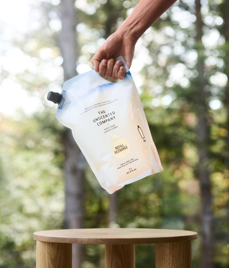 Une main tient une pochette recharge de 2 L de shampoing quotidien The Unscented Company au-dessus d’une petite table en bois, avec une forêt en arrière-plan.