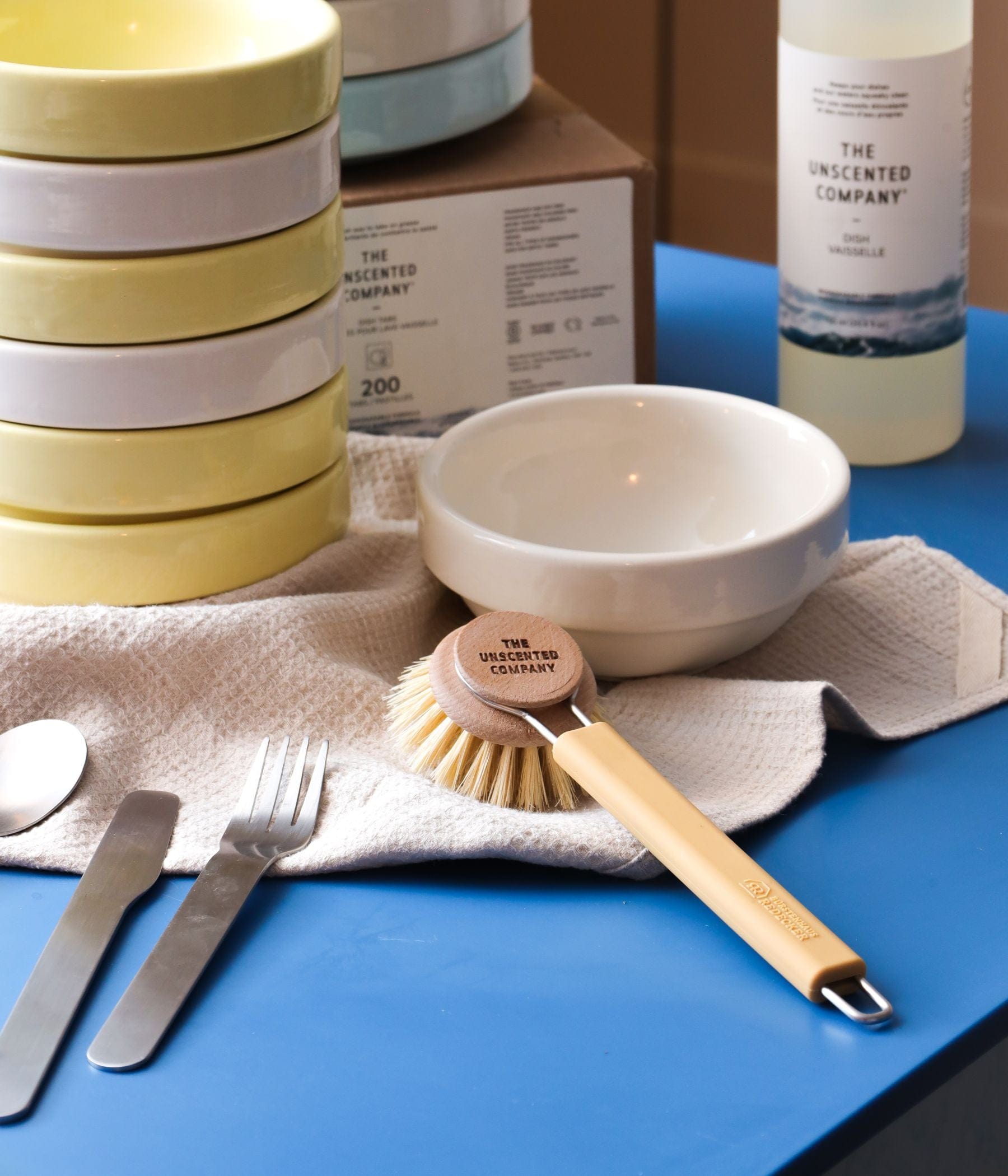 The Silicone Dish Brush Handle + Wood Dish Head in Sand next to cutlery and bowls, a 750ml bottle of Dish Soap is in the background.