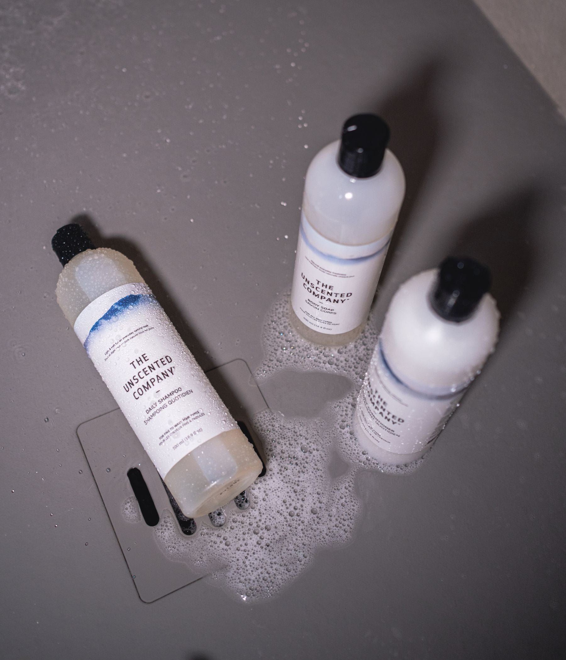 The Unscented Company shampoo, conditioner, and body soap bottles in a shower with bubbles.