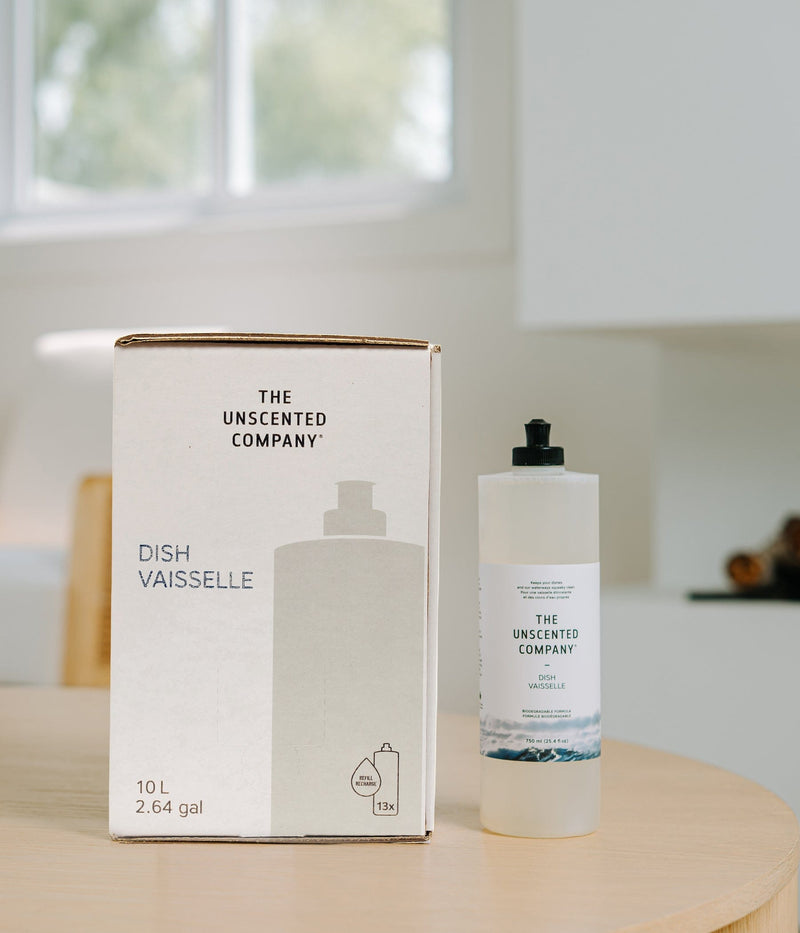 Refill box and bottle of The Unscented Company Dish / Vaisselle dish soap on a wooden table, showing 10 L (2.64 gal) refill packaging with 13x refill icon and 750 ml bottle, photographed in a bright minimalist kitchen.