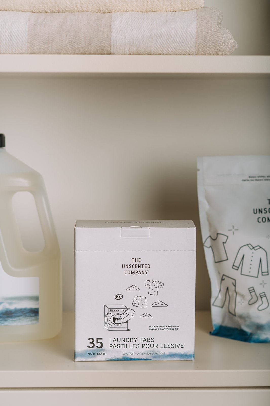 A box of The Unscented Company 35 Laundry Tabs placed on a shelf next to a bottle of liquid detergent and a bag of laundry whitener.