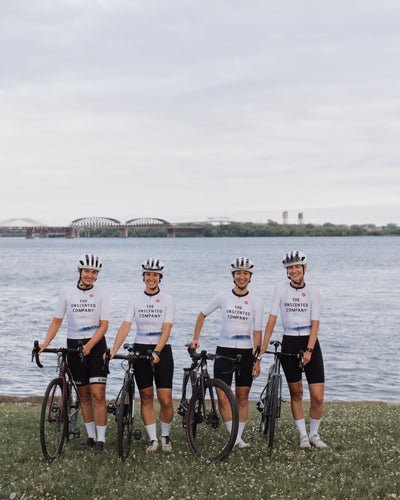 Cyclists wearing The Unscented Company jerseys pose with their road bikes by the water during a group ride, promoting an active lifestyle and sustainable community initiatives.