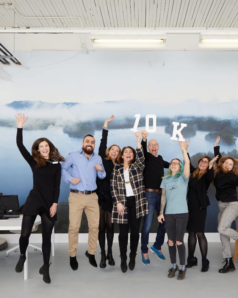 Group of cheerful people jumping and celebrating indoors, holding up letters that spell ‘10K