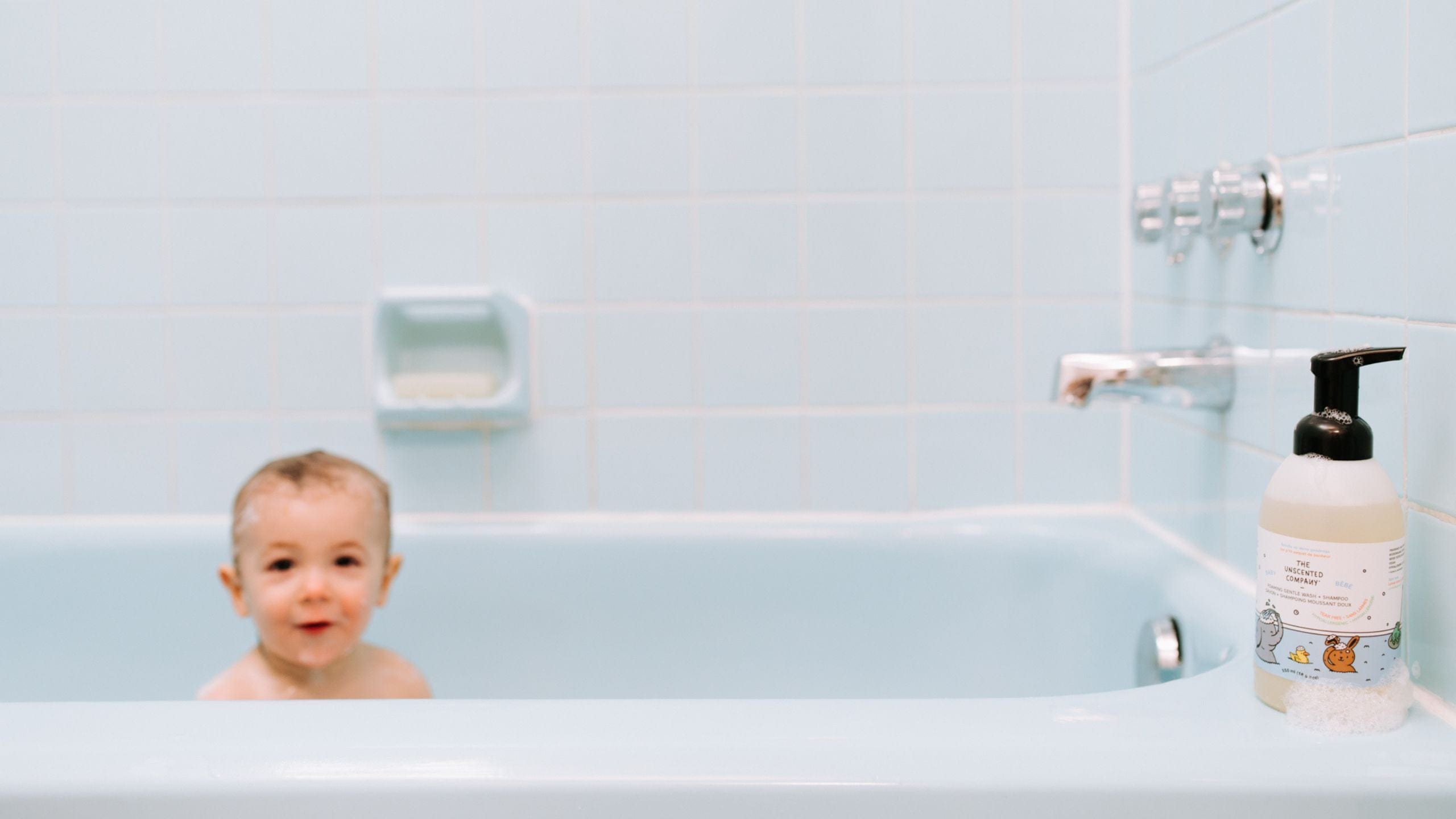 Baby in the bath with a bottle of kids shampoo on the tub’s edge.