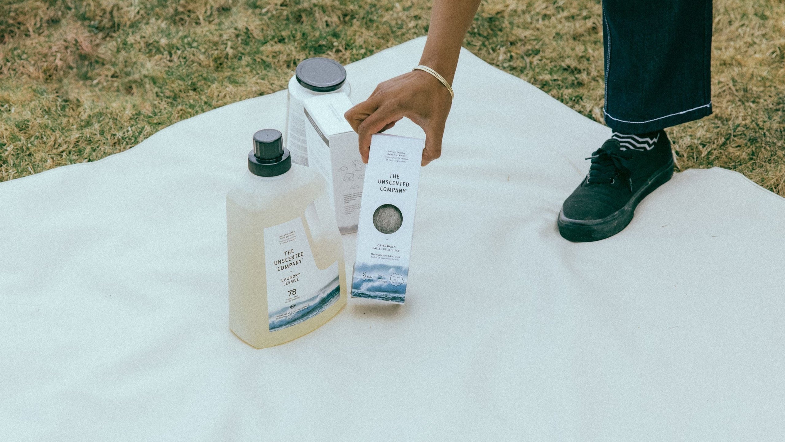 Person picking up The Unscented Company dryer balls outdoors with laundry products on a blanket.