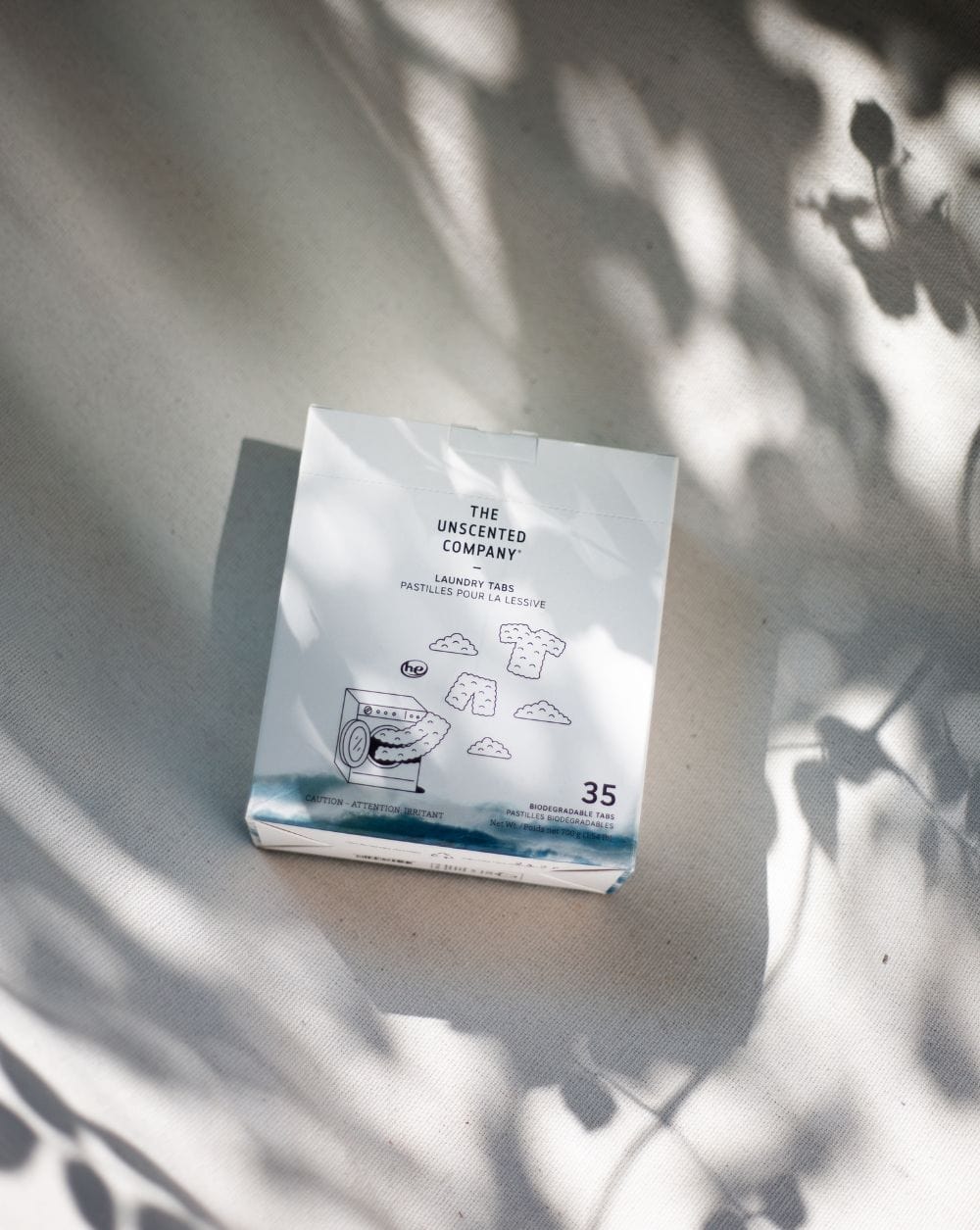 Box of The Unscented Company laundry tabs on a light fabric surface with soft shadows of leaves.