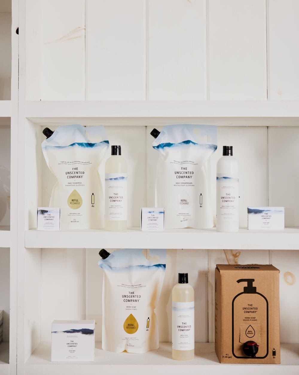 Display of The Unscented Company products on white wooden shelves, including refill pouches, bottles, soap bars, and a cardboard hand soap dispenser.
