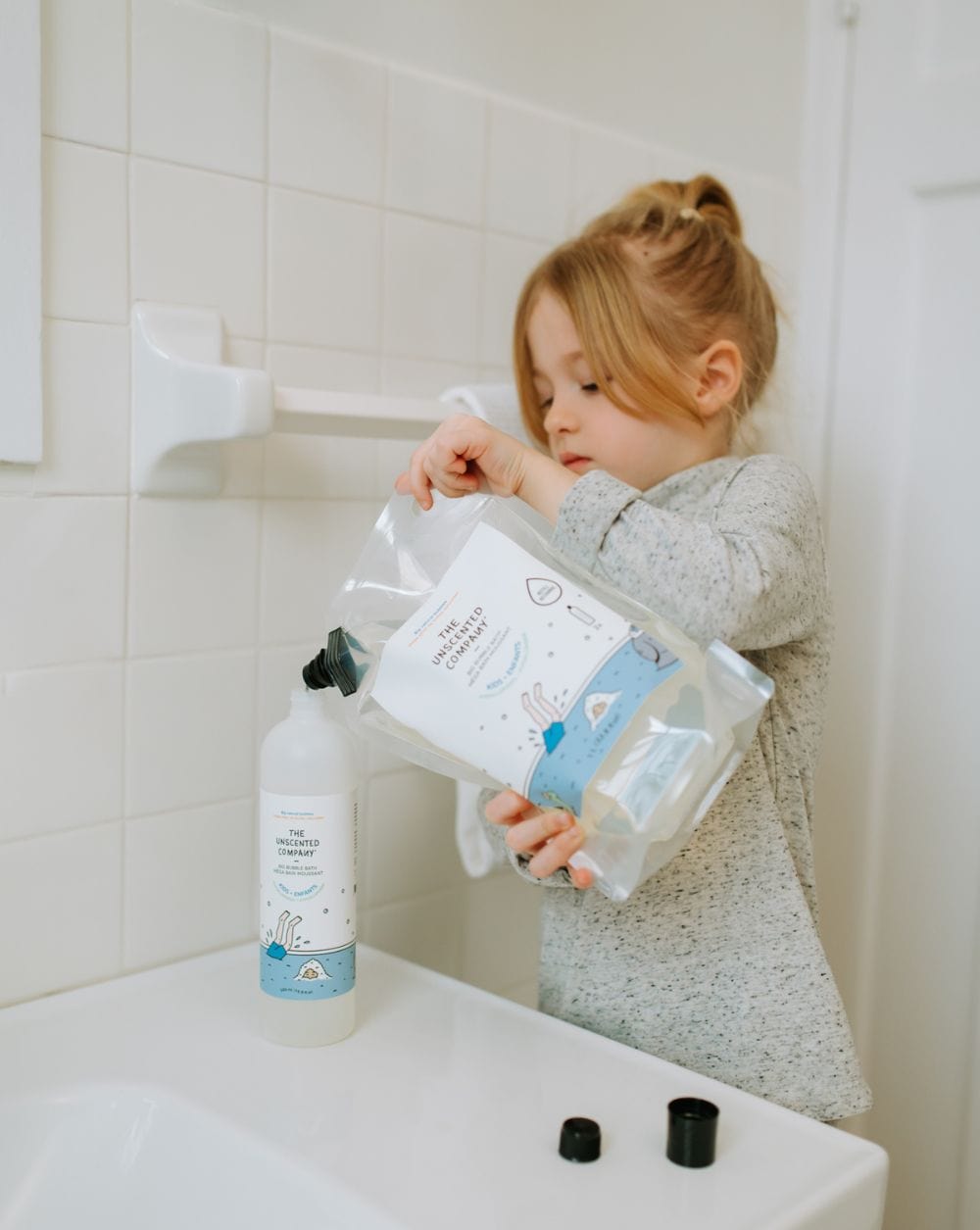 : Child refilling a bottle of The Unscented Company kids shampoo at a sink.
