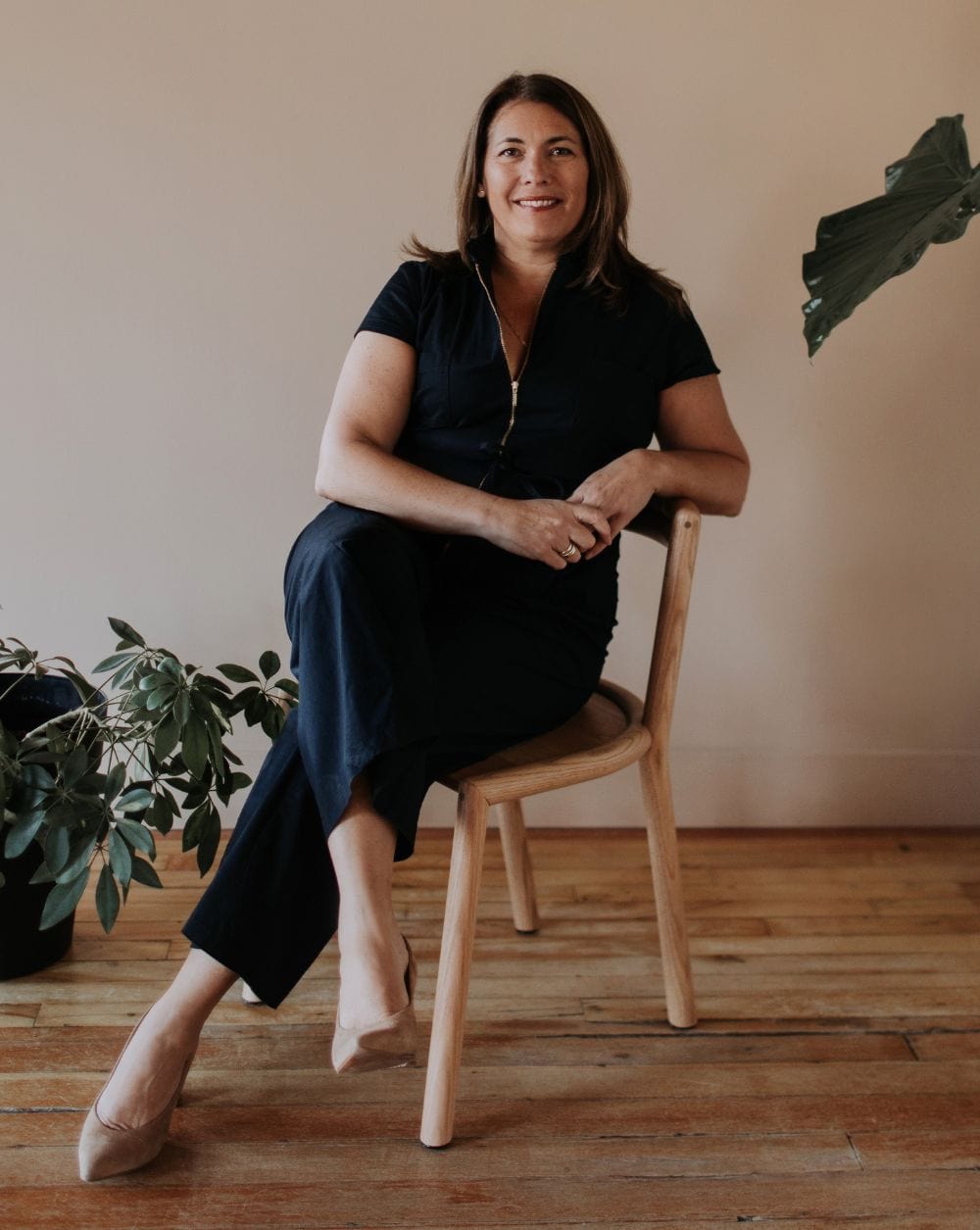 Woman sitting on a wooden chair in a warmly lit room with plants.