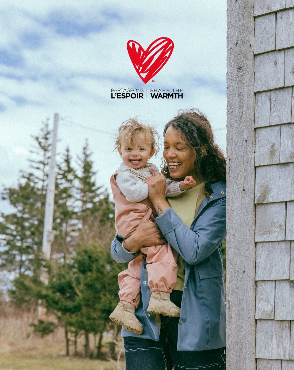 Woman holding a smiling child outdoors under a cloudy sky, next to a wooden building, with the "Share the Warmth" logo above.