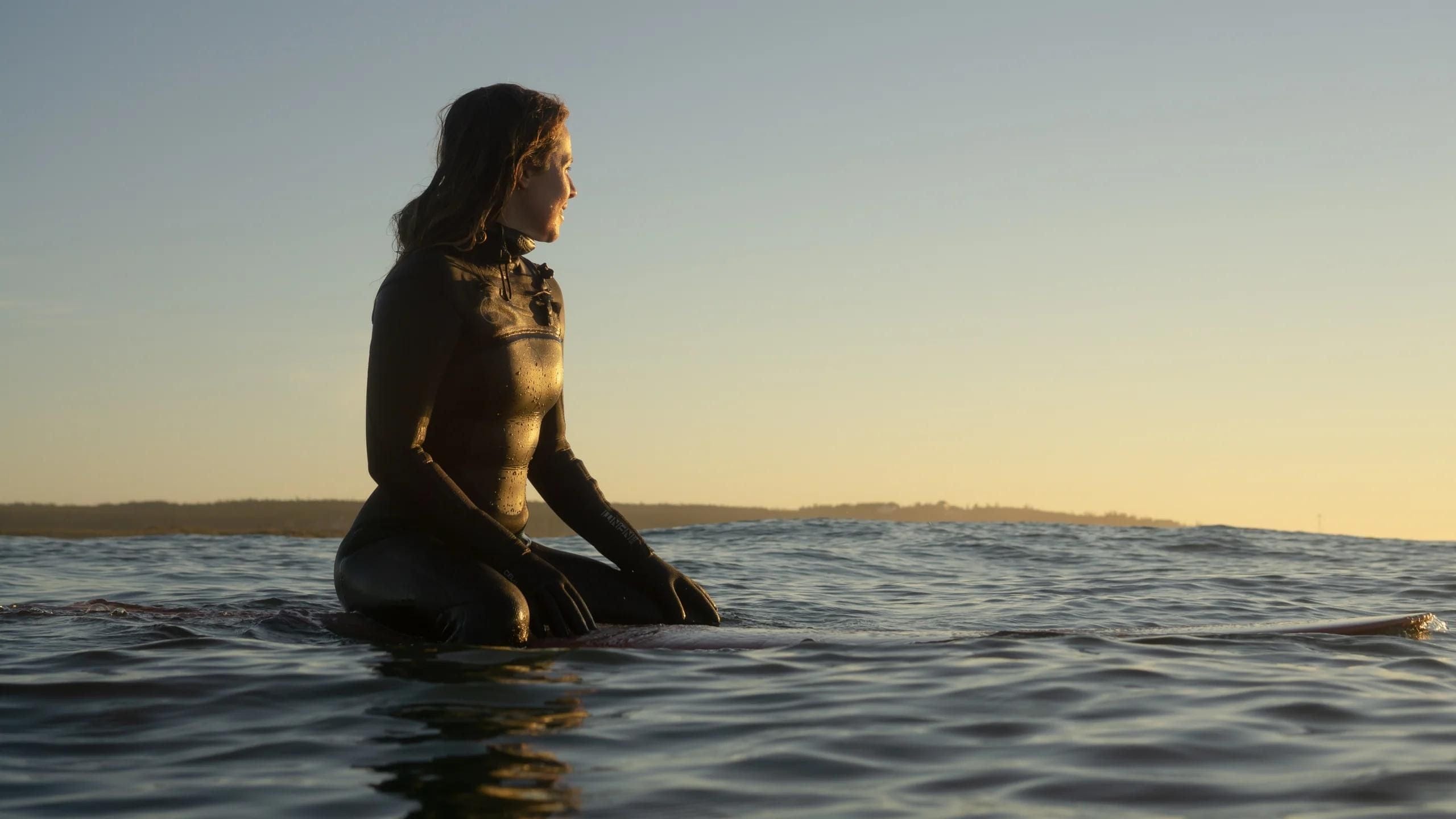 A woman in a wetsuit sits on a surfboard in the ocean at sunset.