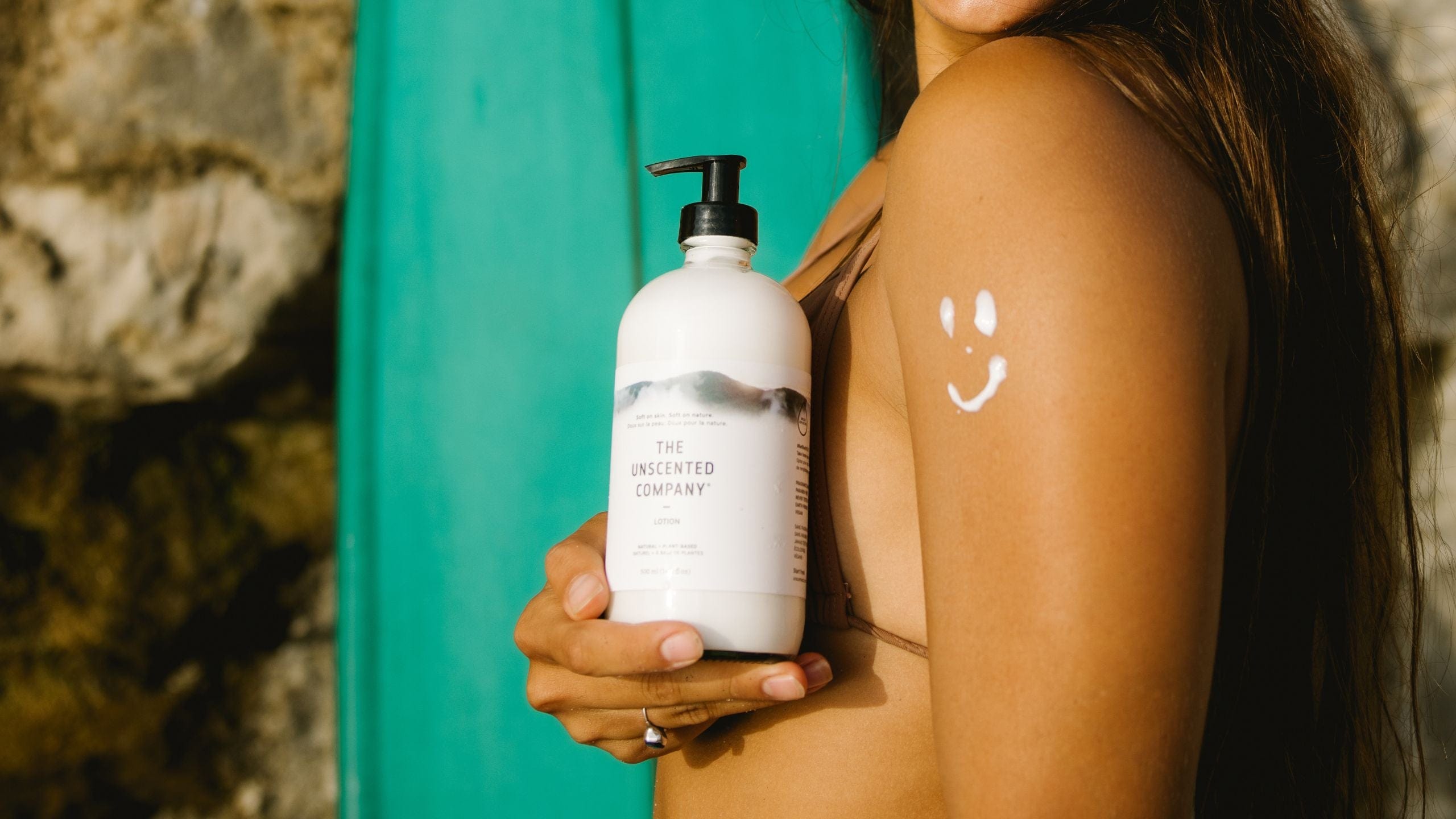 Woman holding a bottle of The Unscented Company lotion with a lotion smiley on her shoulder.