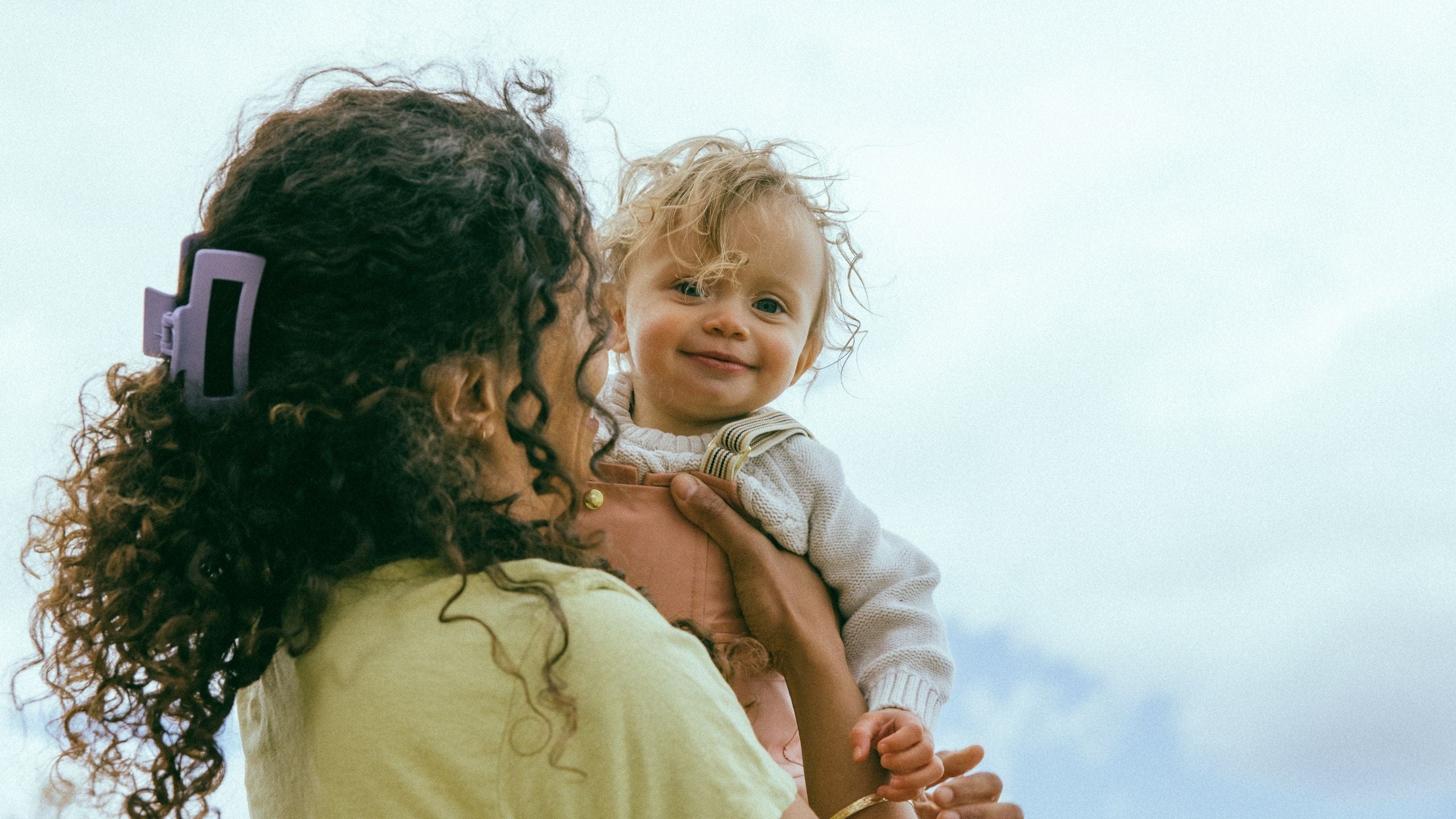 Woman holding a smiling toddler outdoors under a cloudy sky.