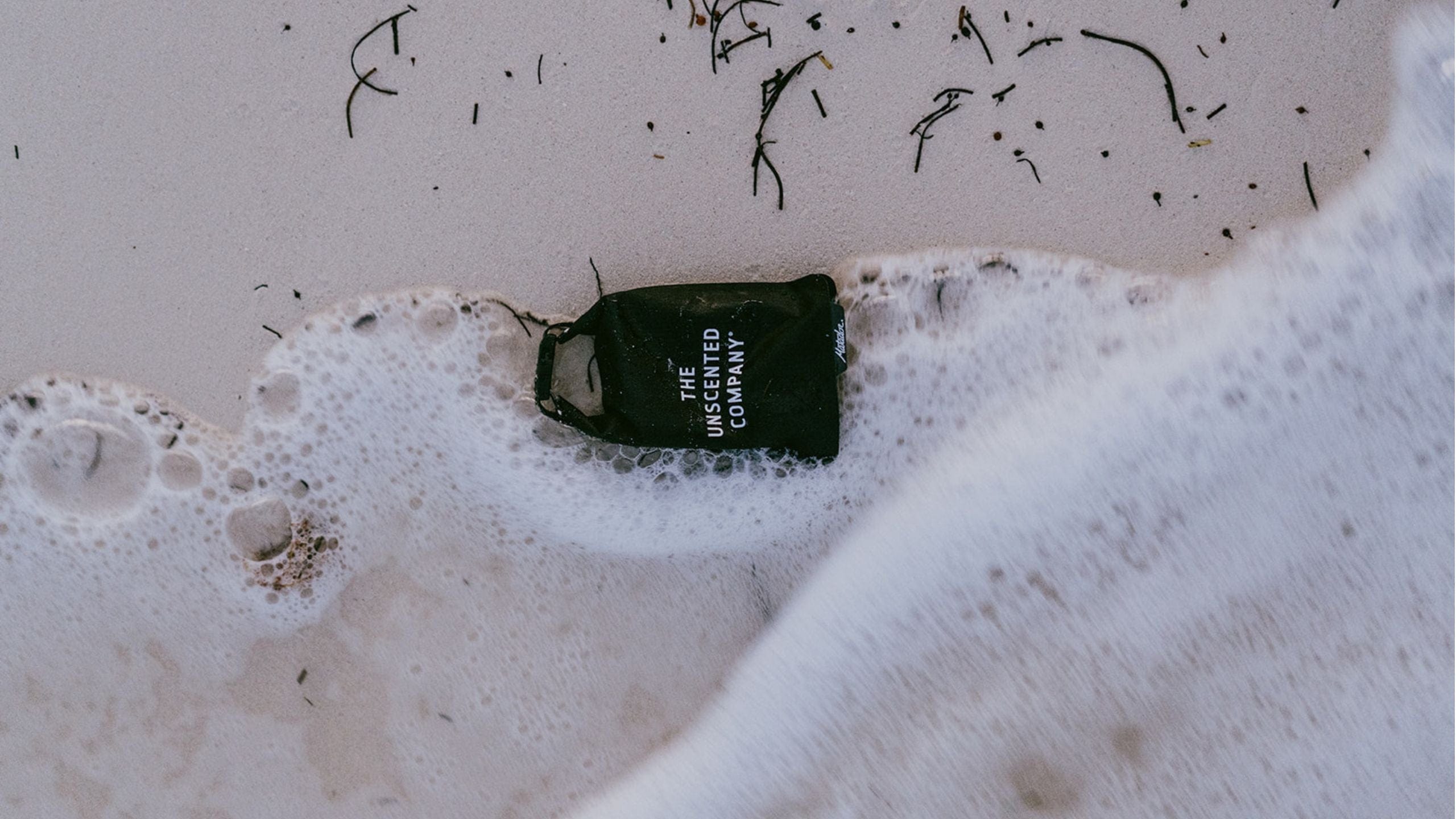 The Unscented Company bar soap in black wrapping washed by ocean waves on the sand.