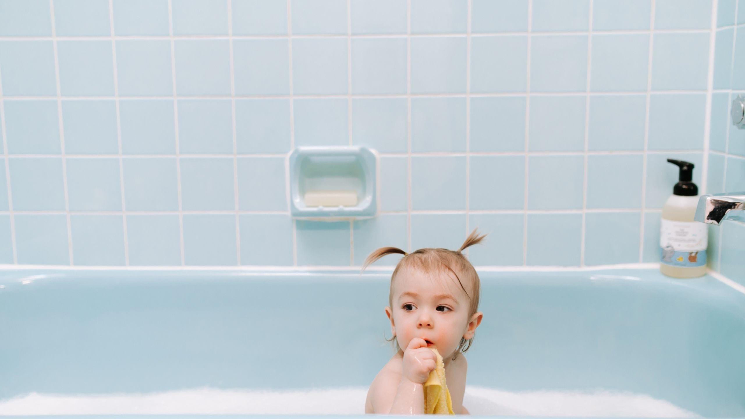 Baby in a blue bathtub holding a washcloth, with The Unscented Company kids shampoo bottle on the edge.