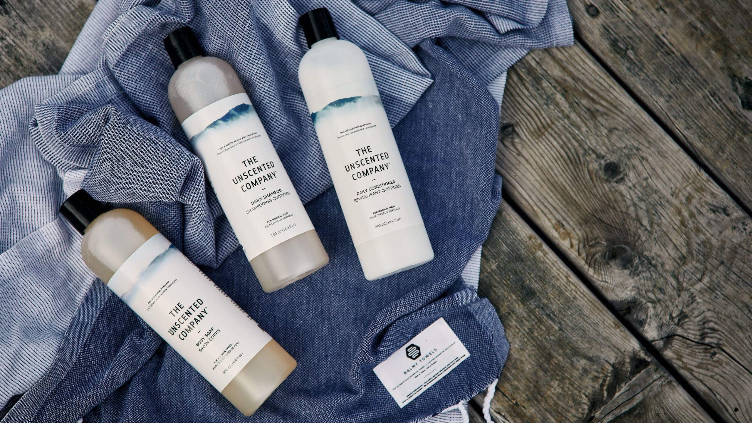 Three bottles of The Unscented Company products (body soap, shampoo, conditioner) arranged on a blue towel on a wooden surface.