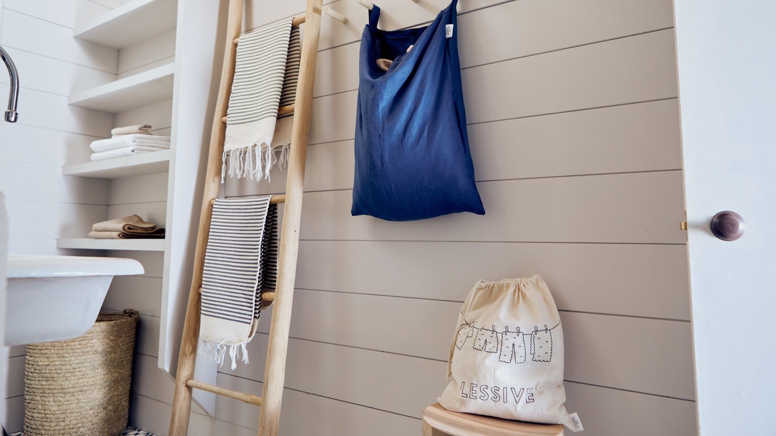 Beige laundry bag labeled “LESSIVE” on a wooden stool next to a ladder with striped towels and shelves with folded linens.